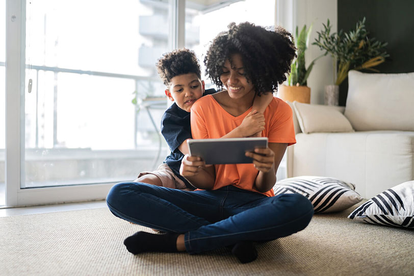 Smiling woman sitting on the floor with a young boy leaning on her shoulder, both looking at a tablet together in a bright living room.